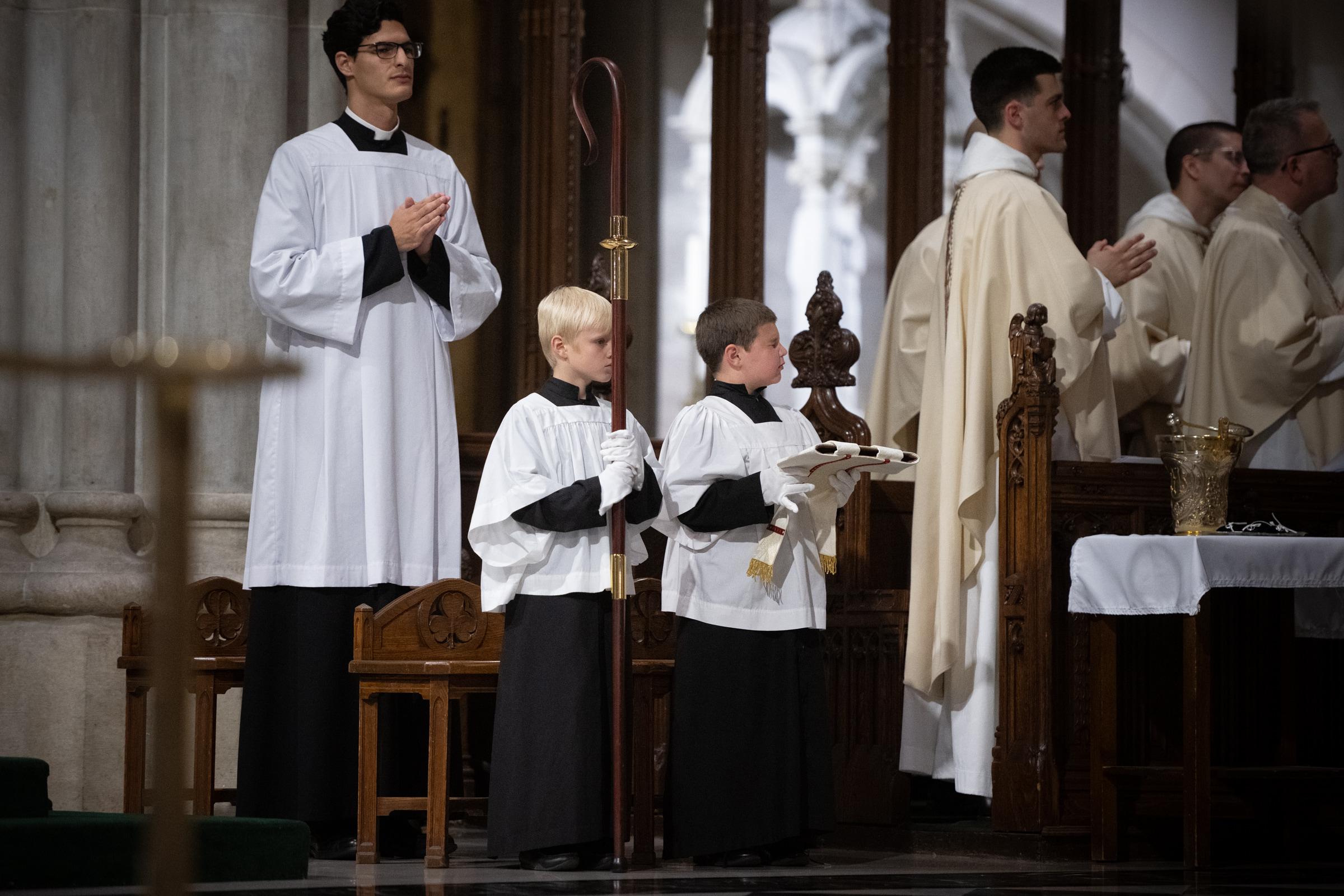 Altar servers hold the crozier and mitre beside clergy in the sanctuary.
