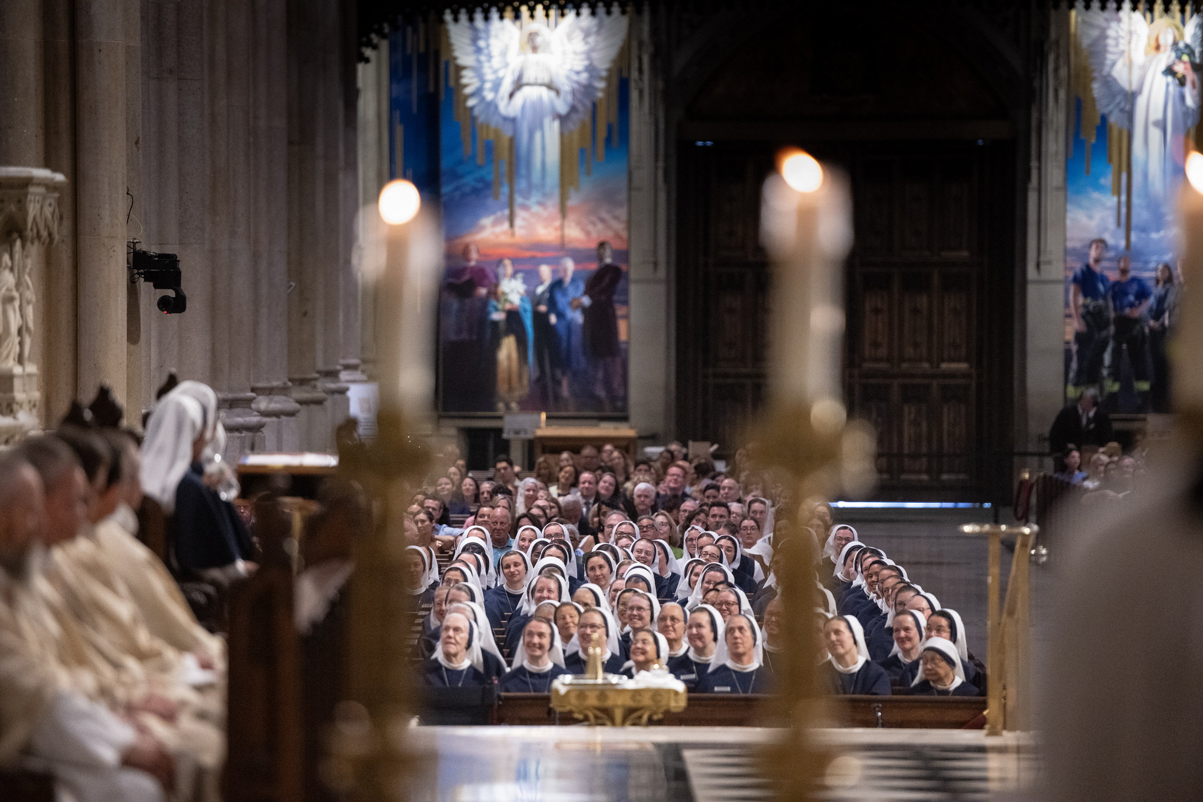 Rows of Sisters listen during Mass, framed through out-of-focus sanctuary candles.