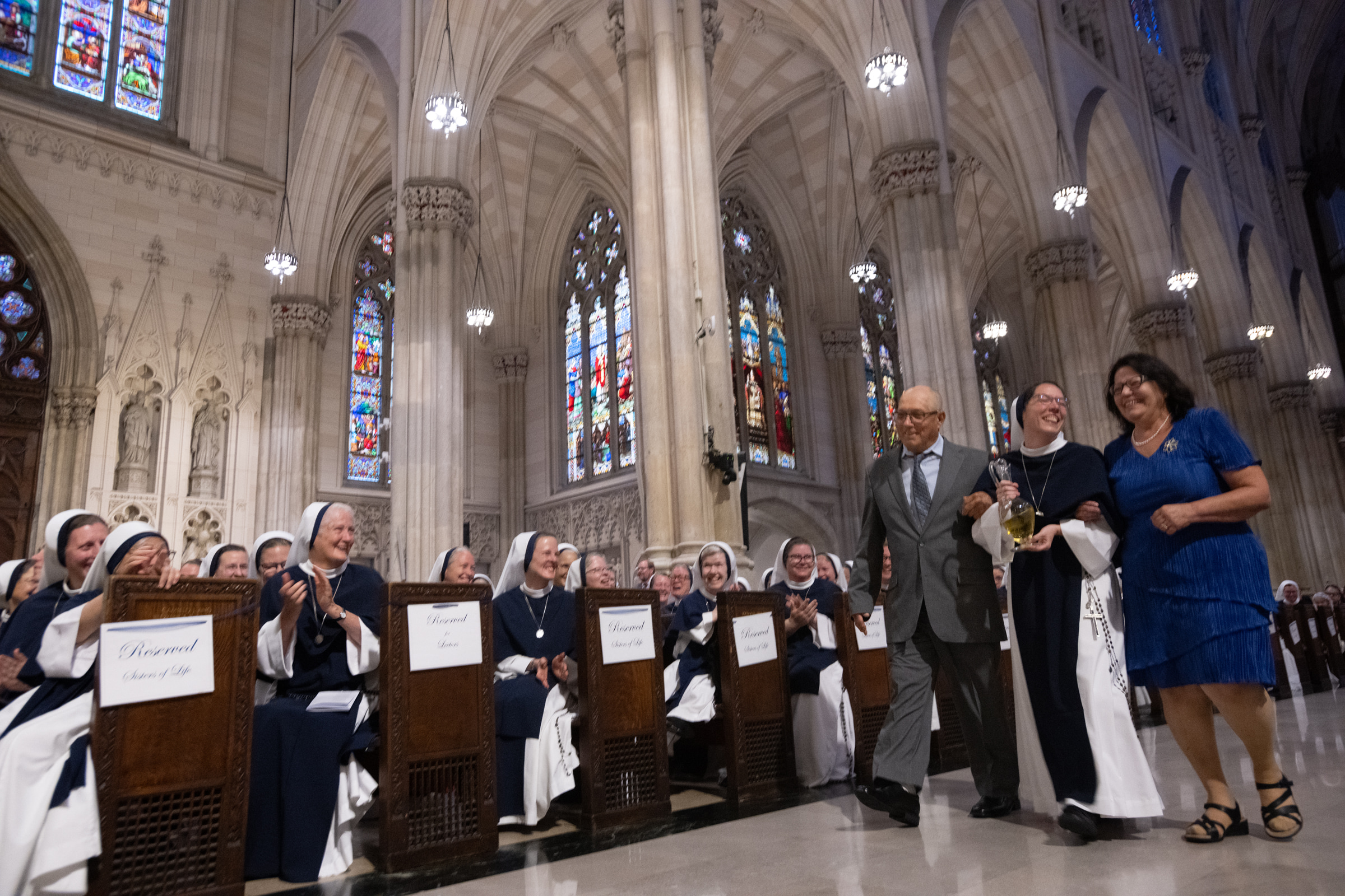Sister Eden Marie, S.V., carries a glass cruet with two guests as Sisters applaud from the pews.