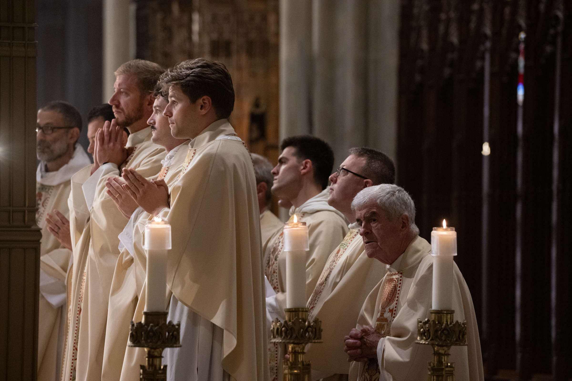 Priests pray behind a row of lit altar candles.